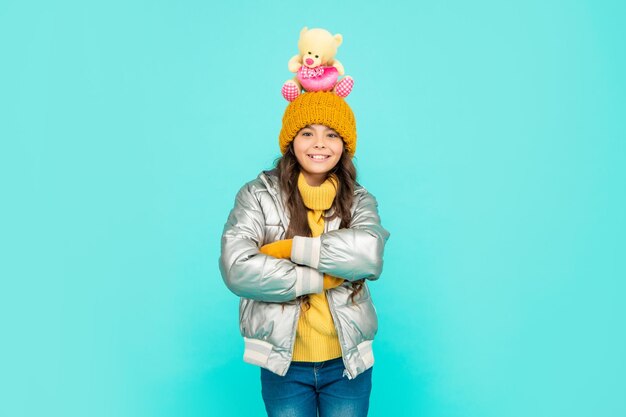 Photo happy teen girl with toy bear crossed hands. child wear warm clothes on blue background. express positive emotion. winter fashion. love toy for valentines day. kid in puffer jacket and hat. childhood.