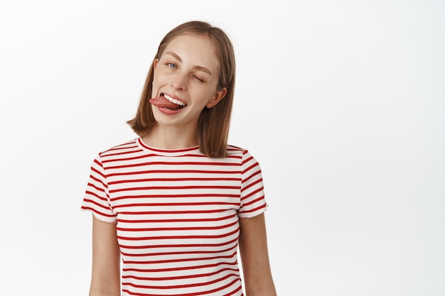 Happy teen woman with perfect teeth, showing her white smile and tongue, winking at front cheerful, standing in striped t-shirt, recommend dental clinic, white wall