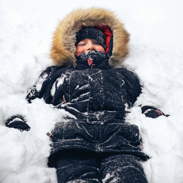 Happy teenage boy dressed in warm winter jacket with fur hood lies in the snow Winter fun