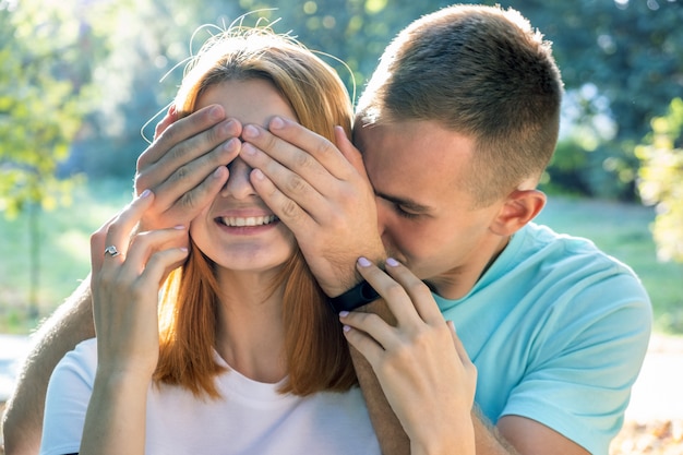 Happy teenage couple having fun together outdoors in sunny summer park surprising one another with closing eyes.