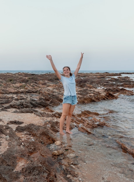 Happy teenage girl standing on cliff by the sea at sunset wearing light blue t-shirt, jeans shorts and holding her hands up. T-shirt mockup