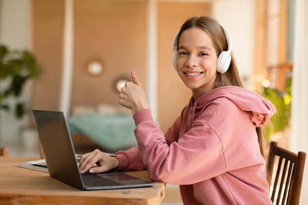 Happy teenage girl studying online using laptop and showing thumb up wearing headphones and smiling at camera