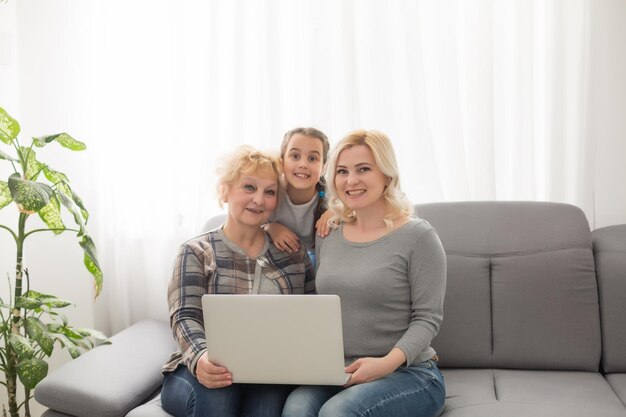 Happy three generations of women sit relax on couch laugh watching funny video on laptop, smiling positive females grandmother, mother and daughter have fun rest on sofa enjoy movie on computer