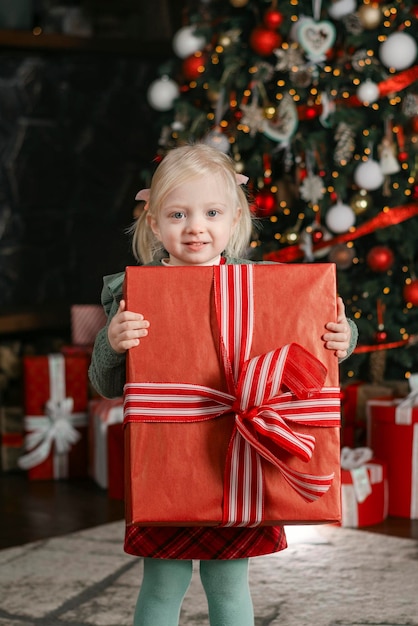 Happy threeyears old girl holds large gift box and looks into camera on xmas tree background Christmas Eve Vertical frame