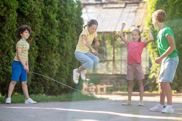 Happy time. Joyful emotional girl with pigtails jumping rope with friends in green park on sunny day