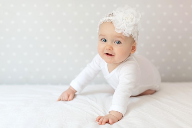 Happy toddler baby girl in a white bodysuit sitting on bed at home and looking at the camera with smile. Copy space for text