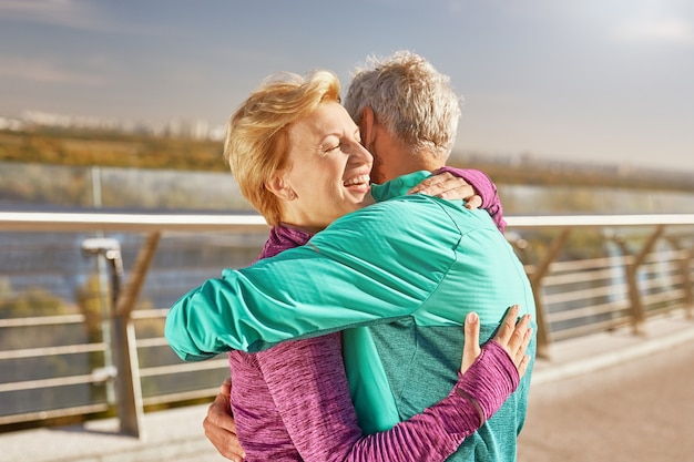 Happy together active mature family couple in sportswear embracing after having workout in the city