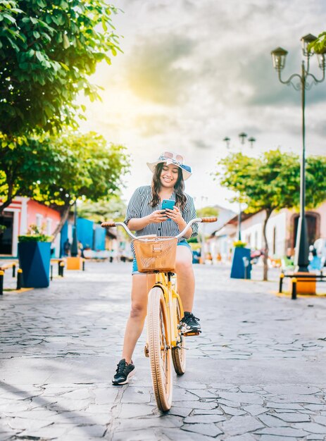 Happy tourist girl on bicycle using cell phone on the street Beautiful girl in hat on bicycle with cell phone on the street of La Calzada Granada Nicaragua