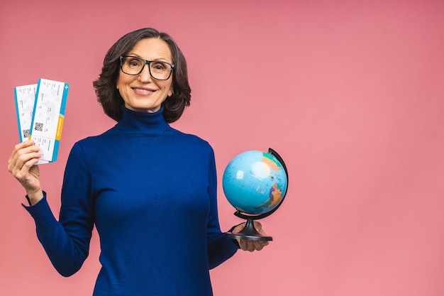 Happy traveler tourist elderly mature senior woman holding tickets and globe isolated on pink background studio. Passenger travel abroad on weekends. Air flight journey concept.