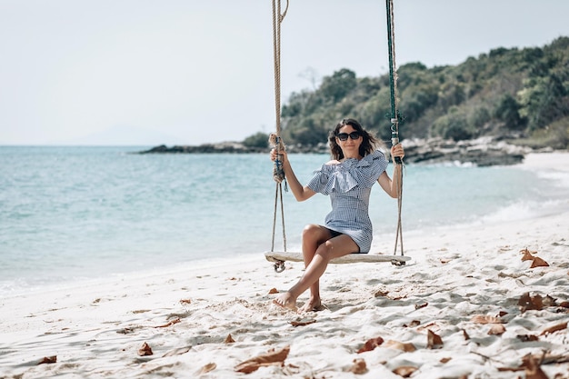 Happy traveler woman in white and black plaid dress  and black sunglasses relaxing on swing. Tourist sea beach Thailand, Asia, Summer holiday vacation travel trip. Phuket. Thailand