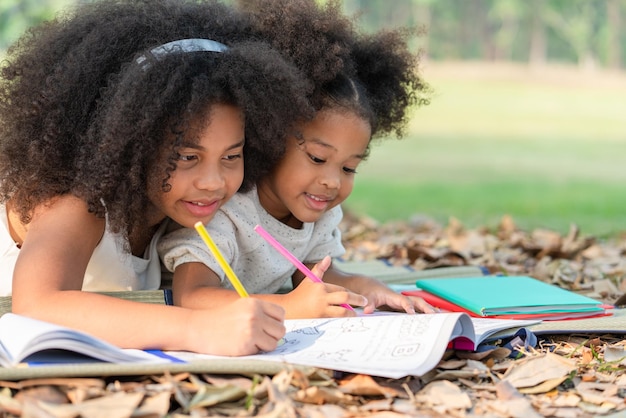 Happy two African American little girls lying drawing in the coloring book for kids in park