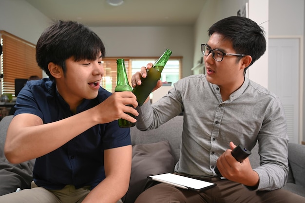 Happy two asian man clinking their glasses of beer while cheering football match on sofa