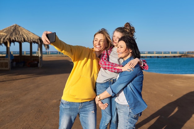 Happy two middle aged women and girl child taking selfie on smartphone sea beach background