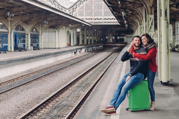 Happy unshaven husband and wife embrace at railway station, going to have trip, have vacation.