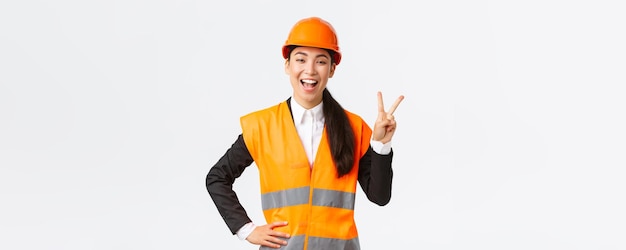 Happy upbeat asian female asian architect in safety helmet and reflective jacket showing peace sign