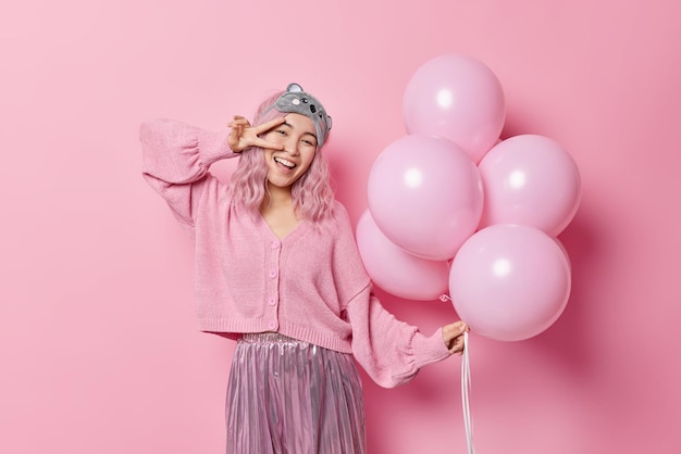 Happy upbeat Asian woman makes peace gesture over eye smiles gladfully poses with inflated balloons dressed in festive clothes celebrates special occasion on costume party isolated on pink wall