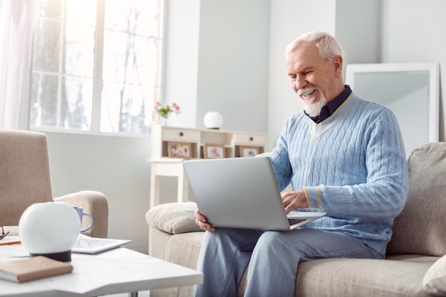 Happy user. Cheerful elderly man sitting on the sofa in the living room and using his laptop, typing a message to his friends