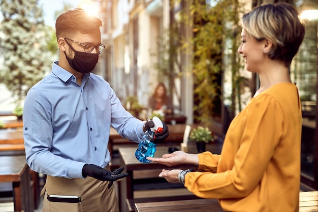 Happy waiter disinfecting hands of a female guest at outdoor cafe