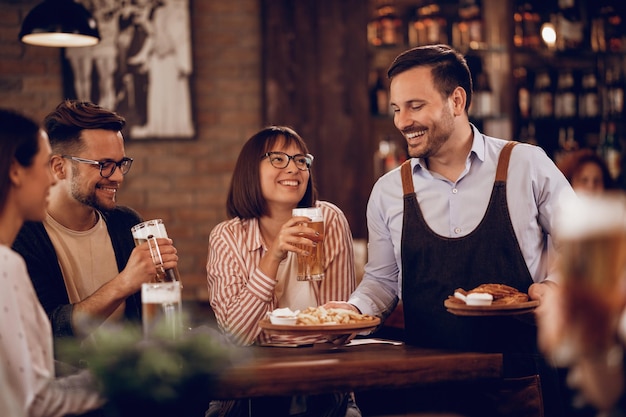 Happy waiter giving food to group of people who are drinking beer in a bar