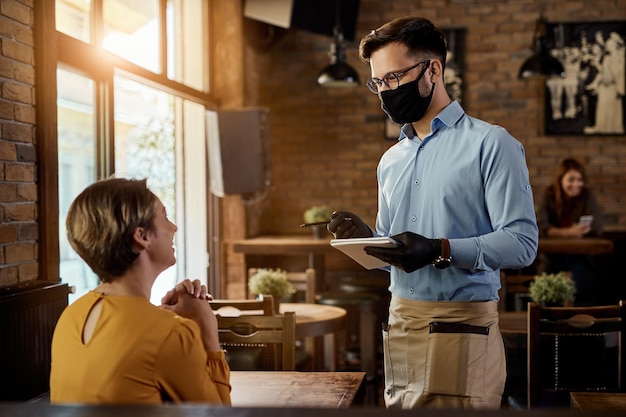 Happy waiter talking to female customer while wearing protective face mask and gloves in a cafe