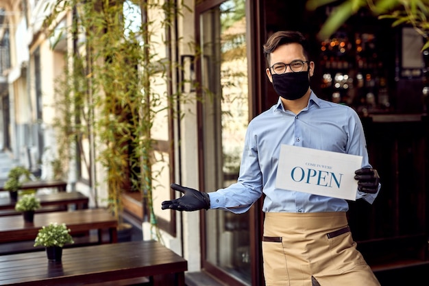 Happy waiter with protective face mask holding open sign while standing at cafe doorway