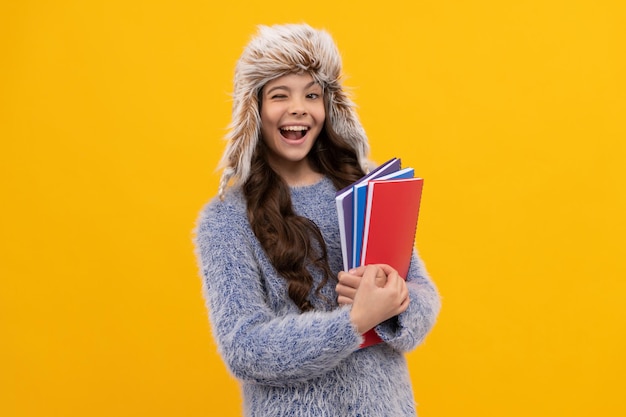 Happy winking kid long hair in hat with copybook on yellow background, knowledge