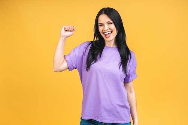 Happy winner Portrait of happy cute young brunette woman with toothy smile raised hands and celebrating victory Isolated over yelow background