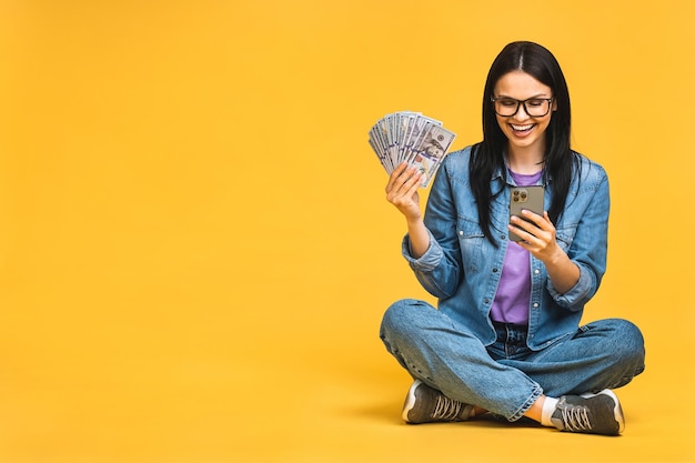 Happy winner Portrait of a smiling cheerful woman using smartphone isolated over yellow background Sitting on the floor in lotus pose holding money bills