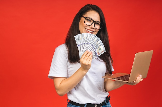 Happy winner Pretty young business woman in casual holding laptop in the office isolated over red background Working with computer holding money bills