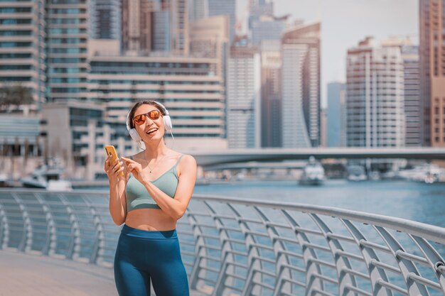 Happy woman in activewear top running along the Dubai Marina embankment and listens to music with headphones and using her smartphone