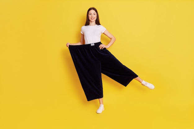 Happy woman after weight loss posing on one hand isolated over yellow wall, wearing white casual t shirt and too big black pants, keeps one hand on hip, rejoices at achieved result.