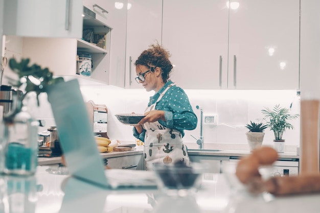 Happy woman alone at home cooking lunch with a smile Ingredients and online computer in foreground defocused Concept of real wife and single lady lifestyle Female people cook in the white kitchen