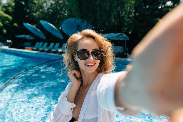 Happy woman blogger is recording live stream near the swimming pool at the hotel A girl in sunglasses on vacation makes a selfie on a mobile phone closeup Vacation and blogging