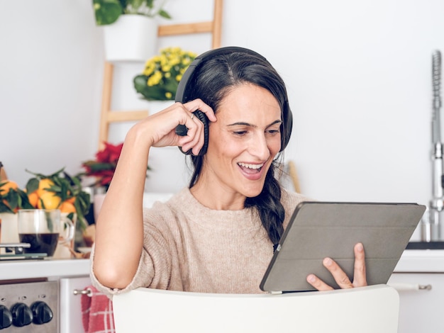 Happy woman blogger in wireless headphone watching video tutorial chat on social media on tablet and smiling while sitting in kitchen