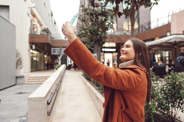 A happy woman in a coat standing on the street and taking a selfie