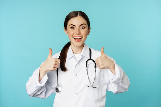 Happy woman doctor medical worker in white coat showing thumbs up in approval like something praise ...