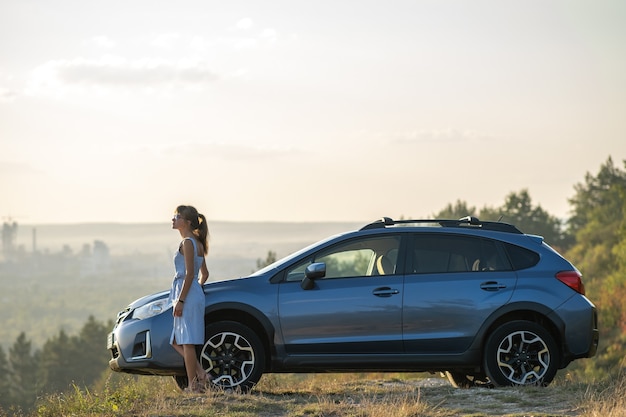 Happy woman driver in blue summer dress enjoying warm evening near her car. Travel and vacations concept.