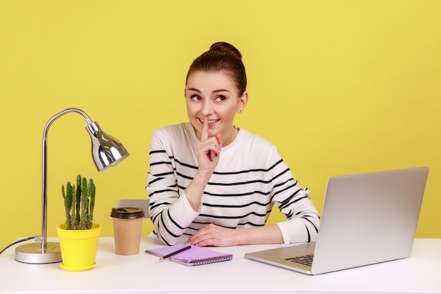 Happy woman employee at workplace shushing with silence gesture asking to keep secret