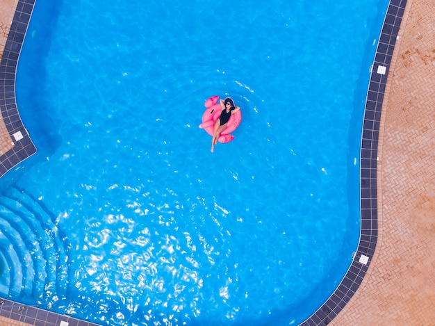 Happy woman on flamingo pool float in pool, drone aerial view