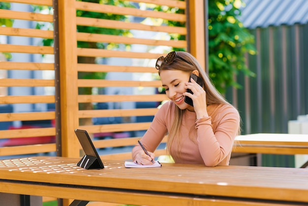 Happy woman freelancer or blogger entrepreneur is sitting at a table in an outdoor cafe and talking on a mobile phone writing notes in a notebook