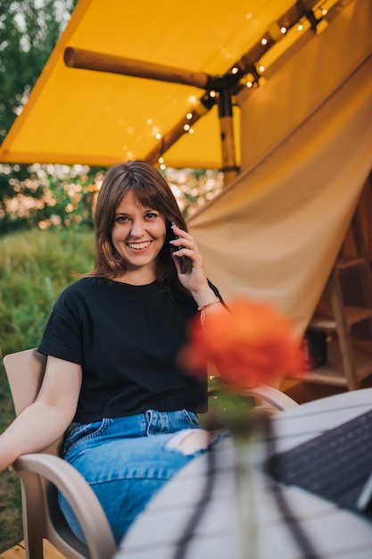 Happy Woman freelancer talking phone and using laptop on cozy glamping tent in a sunny day Luxury camping tent for outdoor summer holiday and vacation Lifestyle concept