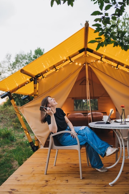 Happy Woman freelancer talking phone and using laptop on cozy glamping tent in a sunny day Luxury camping tent for outdoor summer holiday and vacation Lifestyle concept