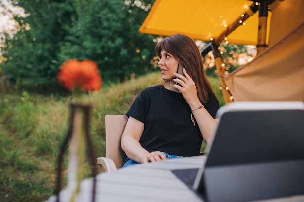 Happy Woman freelancer talking phone and using laptop on cozy glamping tent in a sunny day Luxury camping tent for outdoor summer holiday and vacation Lifestyle concept