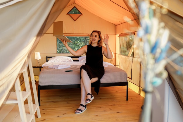 Happy Woman freelancer talking with friends via video call while relaxing on cozy glamping tent in sunny day Luxury camping tent for outdoor summer holiday and vacation Lifestyle concept