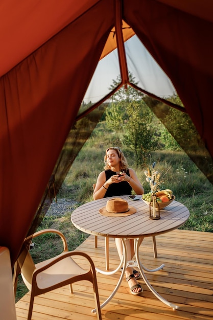 Happy Woman freelancer use phone and drinking cocktail call while relaxing on cozy glamping tent in sunny day Luxury camping tent for outdoor summer holiday and vacation Lifestyle concept