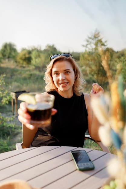 Happy Woman freelancer use phone and drinking cocktail call while relaxing on cozy glamping tent in sunny day Luxury camping tent for outdoor summer holiday and vacation Lifestyle concept