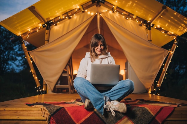 Happy Woman freelancer using a laptop on a cozy glamping tent in a summer evening Luxury camping tent for outdoor holiday and vacation Lifestyle concept