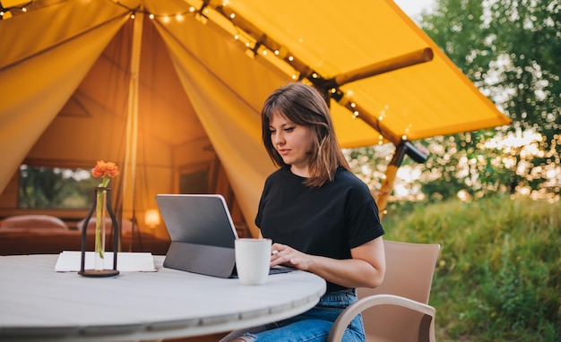 Happy Woman freelancer using a laptop on a cozy glamping tent in a sunny day Luxury camping tent for outdoor summer holiday and vacation Lifestyle concept