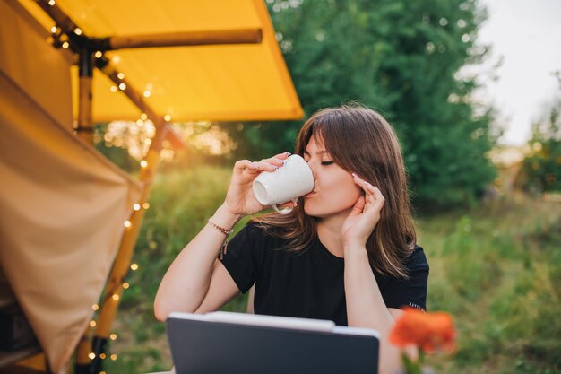 Photo happy woman freelancer using laptop and drink coffee on a cozy glamping tent in a sunny day luxury camping tent for outdoor summer holiday and vacation lifestyle concept