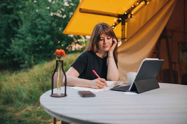 Happy Woman freelancer using laptop and making notes sitting on cozy glamping tent in a sunny day Luxury camping tent for outdoor summer holiday and vacation Lifestyle concept